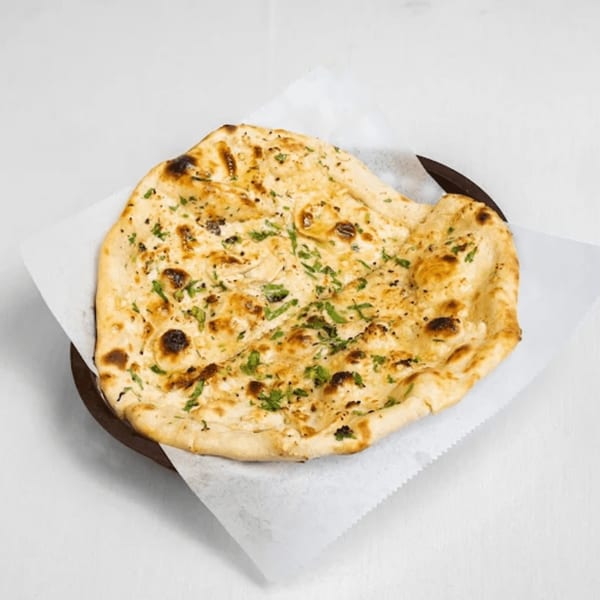 Golden brown naan bread with charred spots and green herbs, served on white paper in a dark bowl.