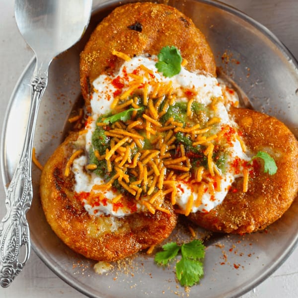 Golden-brown aloo tikki chaat topped with yogurt, green chutney, sev, and spices on a metal plate.