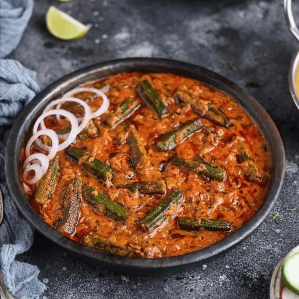 A black bowl filled with a rich, red okra curry, garnished with white onion rings.