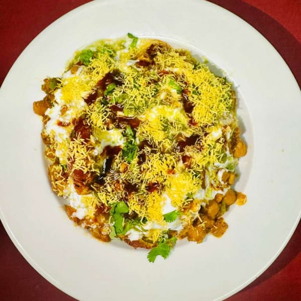 A white plate holds a colorful dish topped with white sauce, brown sauce, green herbs, and yellow crispy noodles.