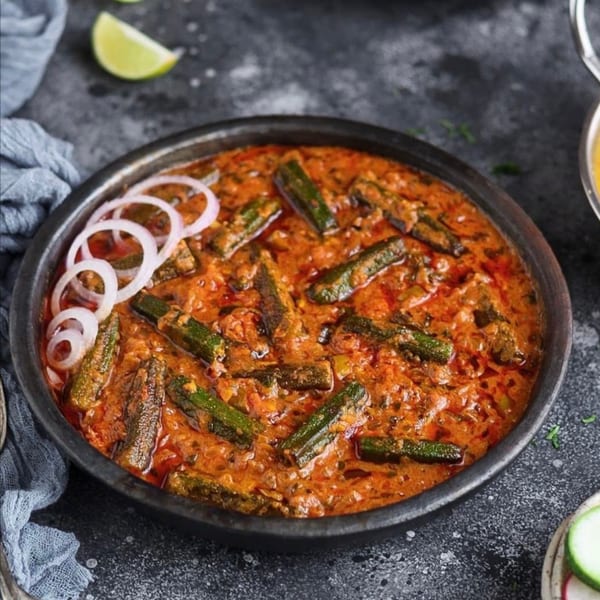 A black bowl of Indian okra curry with red sauce, garnished with white onion rings.